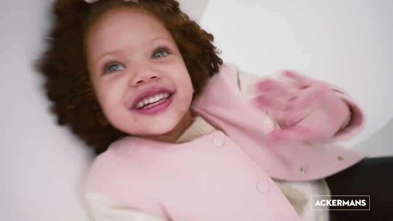 News 126 A young child with curly hair smiles joyfully while waving. The child wears a light pink coat over a cream sweater. The background is softly blurred in white tones. In the bottom right corner, the logo