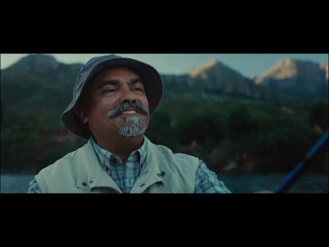 News 120 A middle-aged man with a gray goatee smiles while fishing outdoors. He wears a bucket hat, plaid shirt, and beige vest. Blurred green trees and rugged mountains are visible in the background under a blue sky.