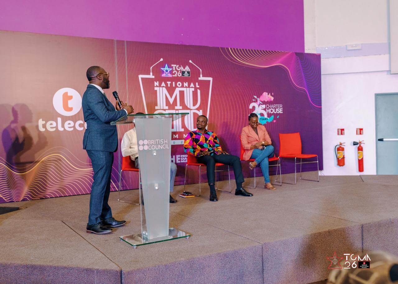 A man stands at a clear podium speaking to two seated men on a stage with a purple and pink backdrop labeled "National Music Summit." Chairs, a fire extinguisher, and event logos are visible. The atmosphere appears formal and professional.