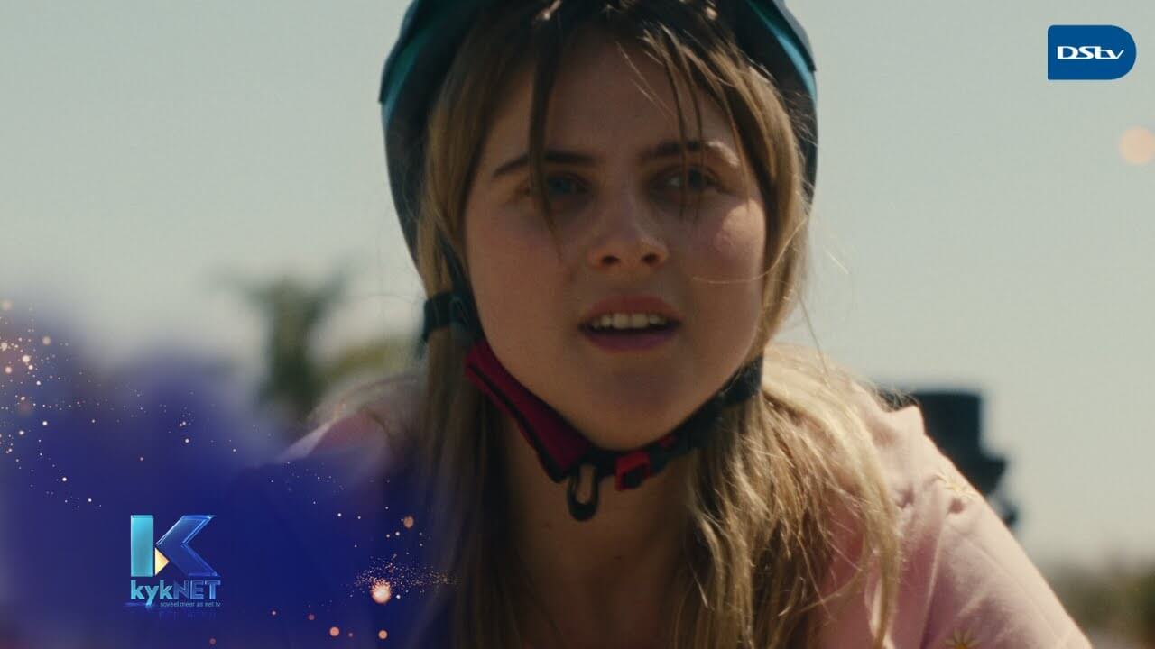 A young woman with long blonde hair wearing a teal helmet looks intently ahead. She is outdoors under sunlight, and part of her face is in shadow. The kykNET and DStv logos appear in the bottom left and top right corners.
