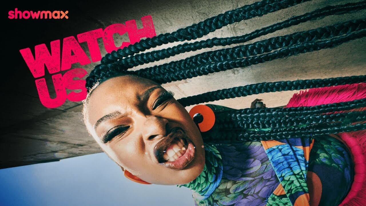 A woman with long braided hair and large orange earrings makes a fierce face at the camera. She wears a colorful, patterned top. Bold pink text reads "WATCH US." The Showmax logo is in the top left corner. The image uses a dynamic, tilted angle.