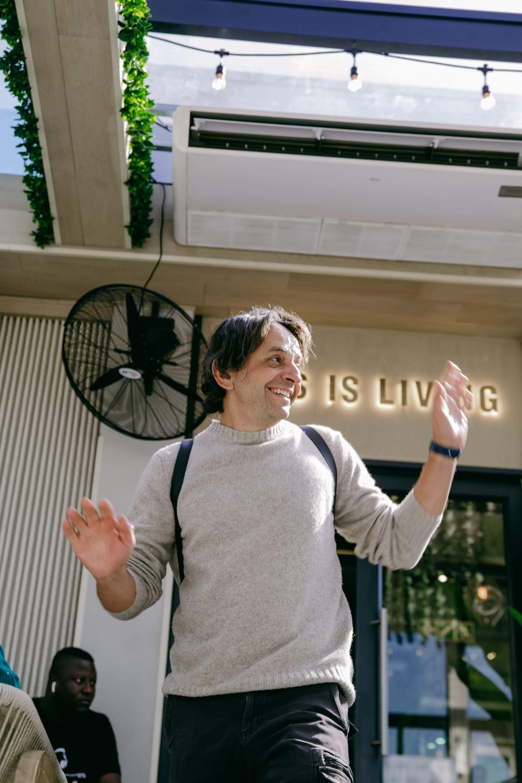 A man with medium-length brown hair, wearing a light gray sweater and black pants, smiles and raises his hands as if mid-dance outside a building. The background shows a large fan, an air conditioner, a few people, and partial text on the wall.