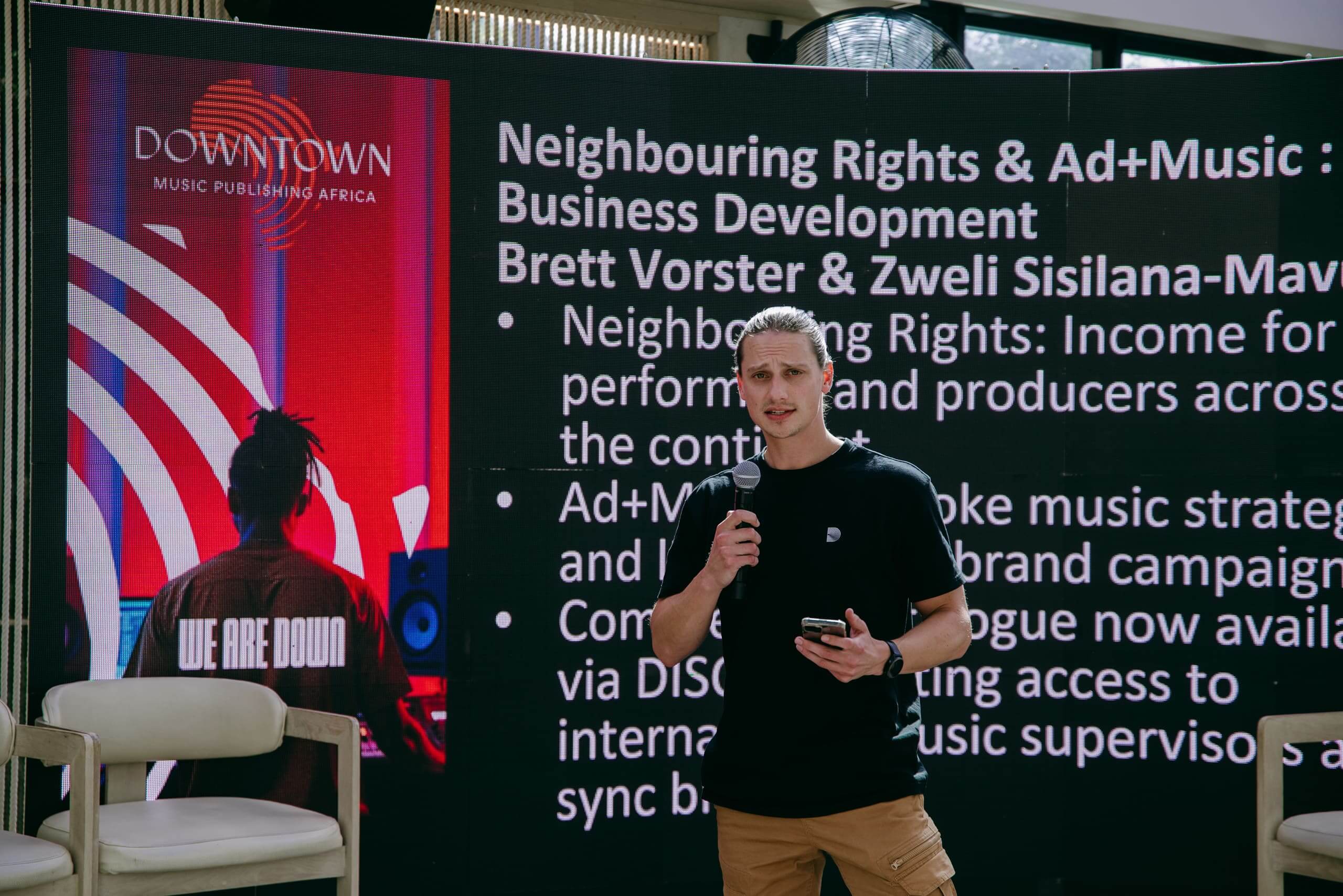 A man with long blond hair in a ponytail speaks into a microphone, holding a phone. He stands in front of a large screen displaying a music business presentation titled 