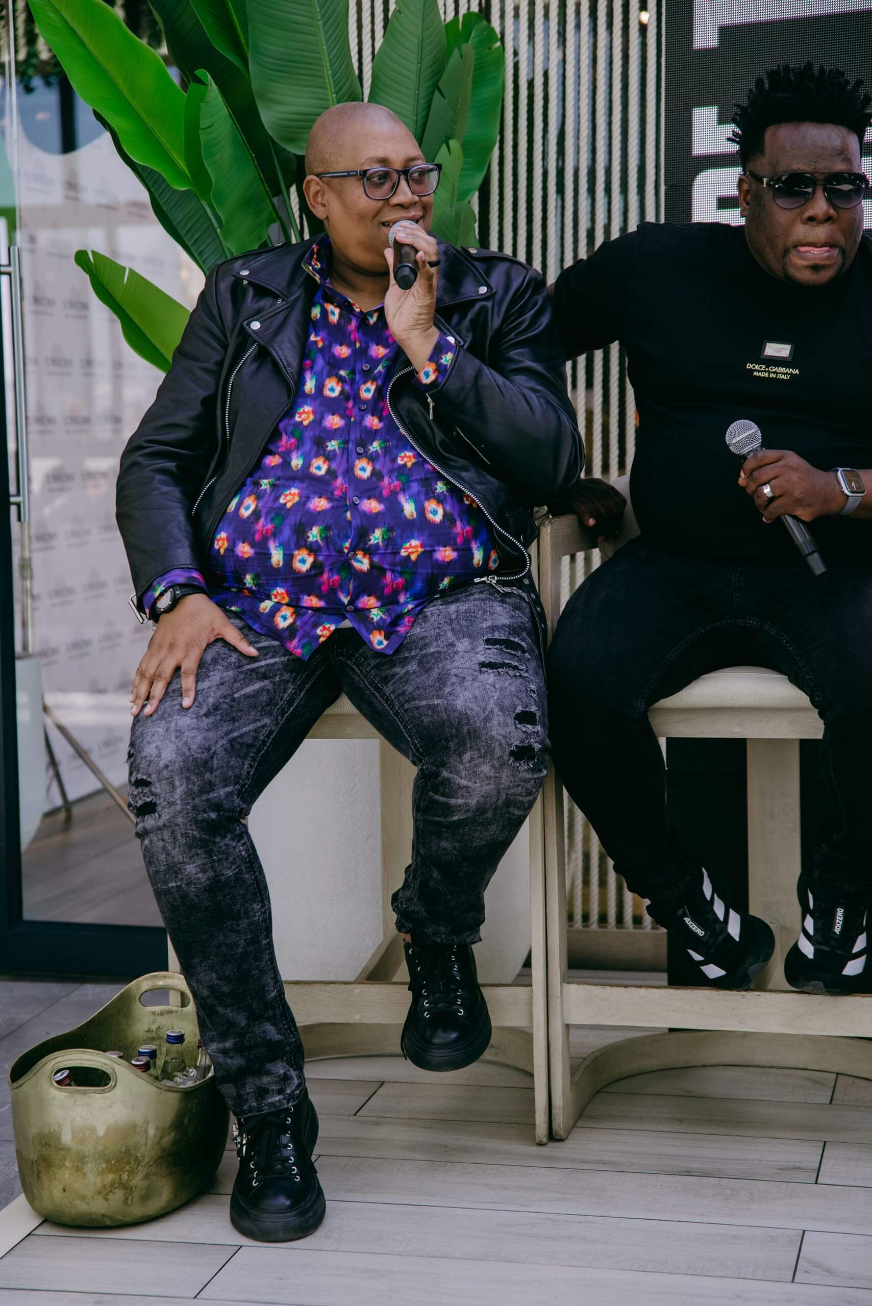 Two men sit side by side on barstools. The man on the left holds a microphone, wears glasses, a black leather jacket, a colorful patterned shirt, ripped gray jeans, and black boots. The man on the right wears sunglasses, a black t-shirt, and dark pants. There’s a large plant and a canister nearby.