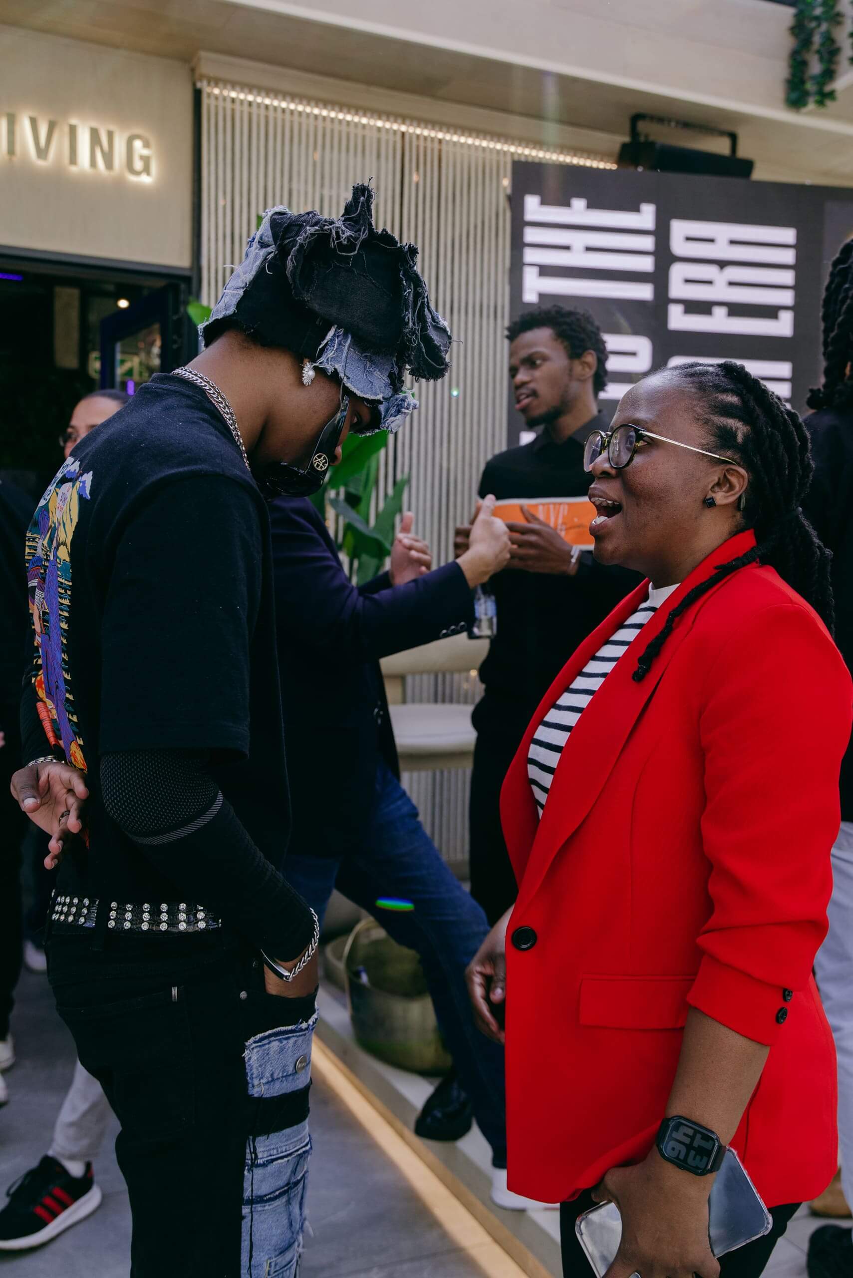 A person in a red blazer and glasses smiles while talking to another person wearing a black shirt and frayed denim hat. In the background, others converse in a modern indoor space with plants and a large black-and-white sign.