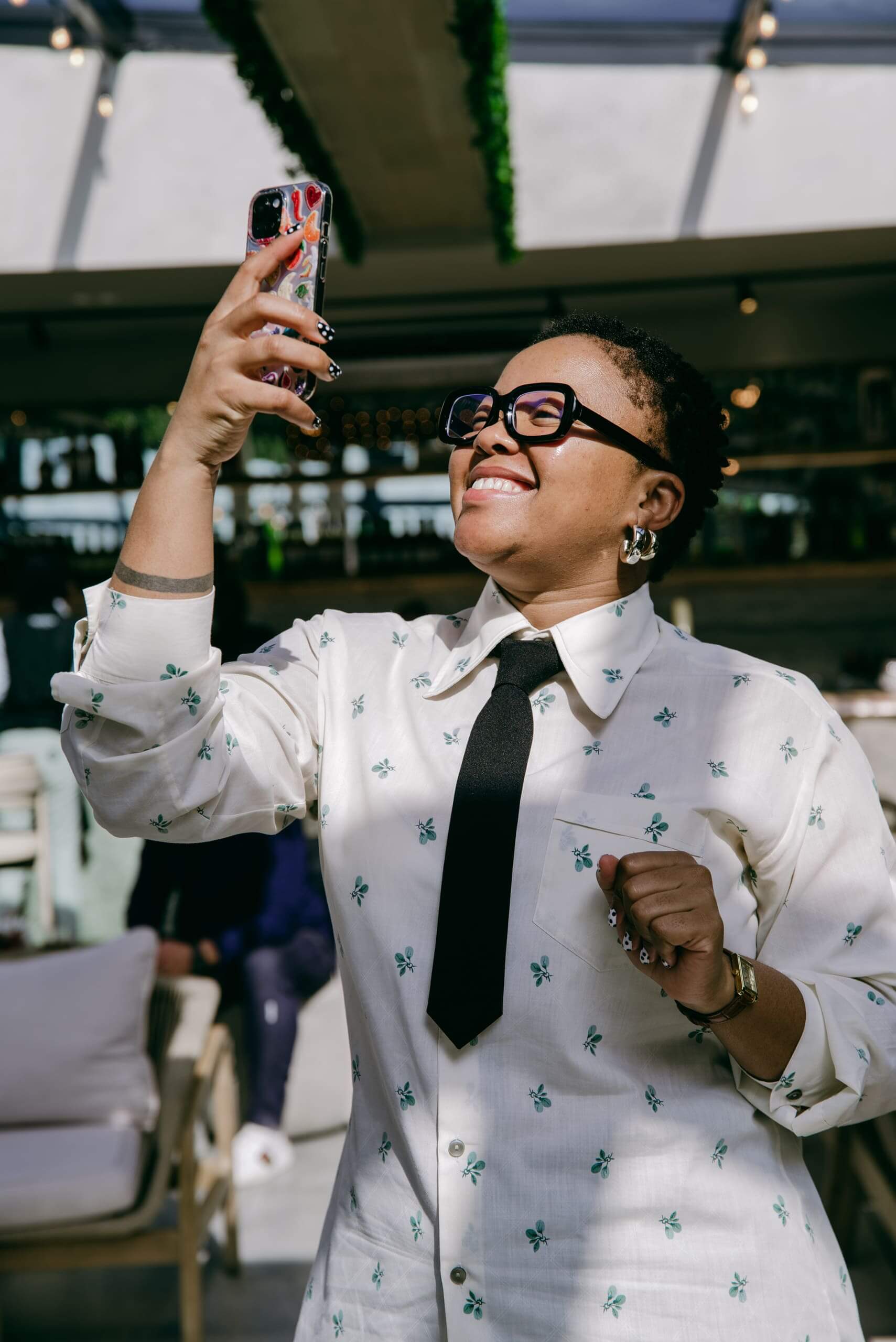 A person wearing glasses, a white shirt with green floral prints, and a black tie smiles while holding a smartphone up, taking a selfie in a bright indoor space with large windows and modern furniture.