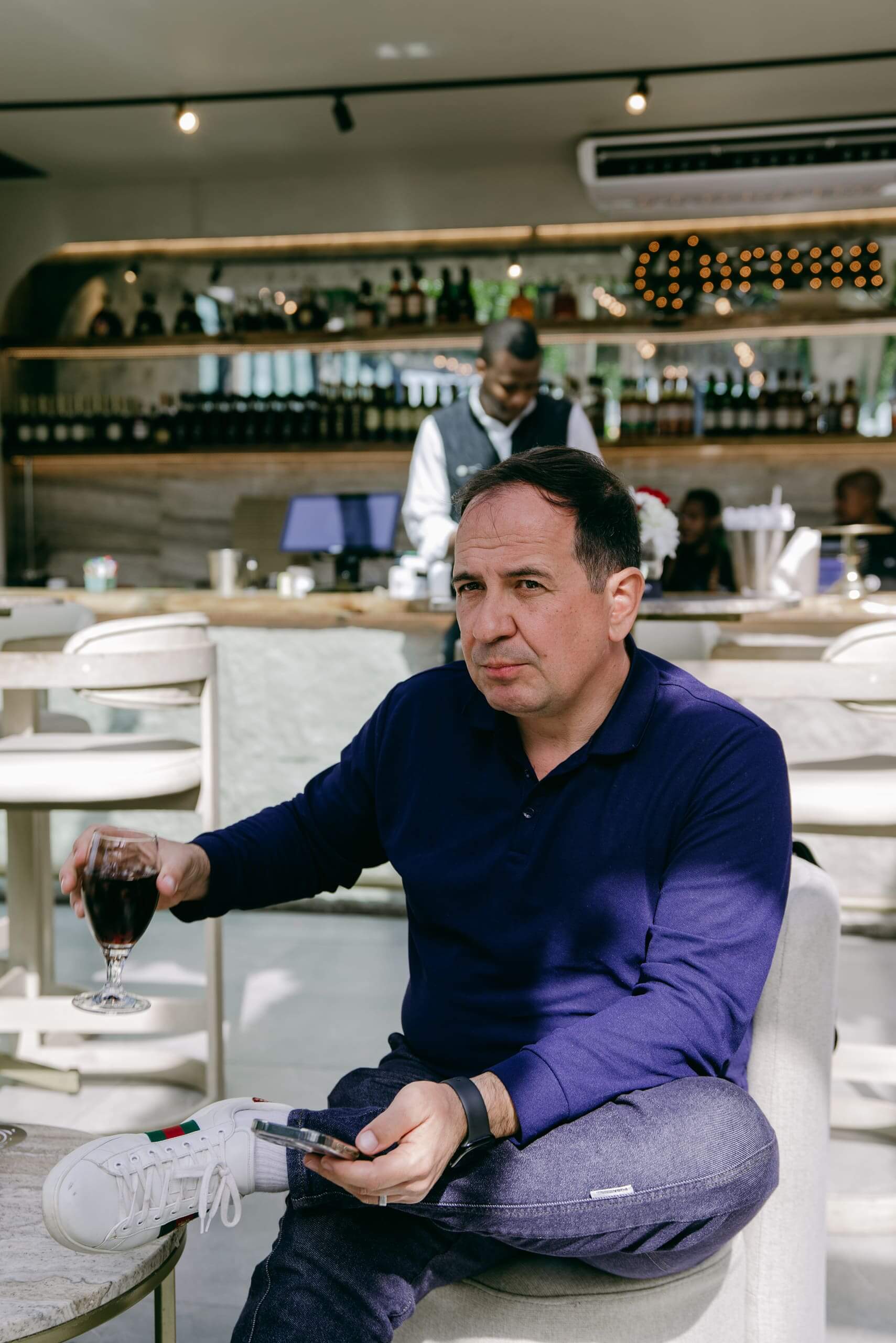 A man in a navy blue shirt and jeans sits in a modern cafe, holding a glass of red wine and a smartphone. He appears thoughtful. Behind him, a bartender works at a counter with bottles and lights. The atmosphere is bright and relaxed.