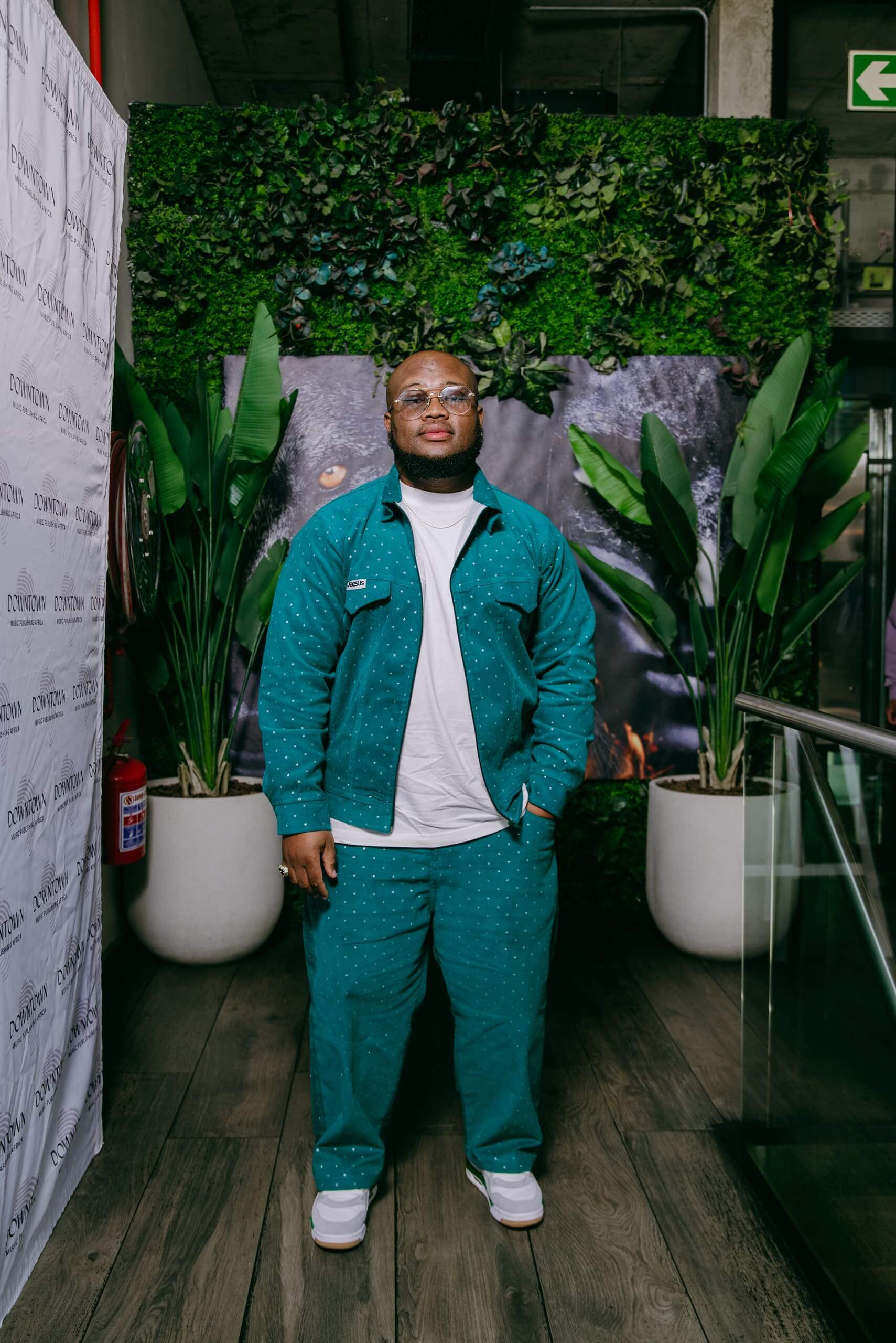 A man stands indoors on a wooden floor, wearing a teal jacket and matching pants with small dots, a white t-shirt, and white sneakers. He has glasses and a beard. Behind him are green plants, a leafy wall, and a partially visible animal mural.