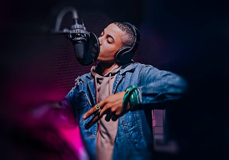 News 112 A young man wearing headphones and a denim jacket raps passionately into a professional microphone in a dimly lit recording studio, gesturing with his hand. The background is dark and blurred, emphasizing his focus and performance.