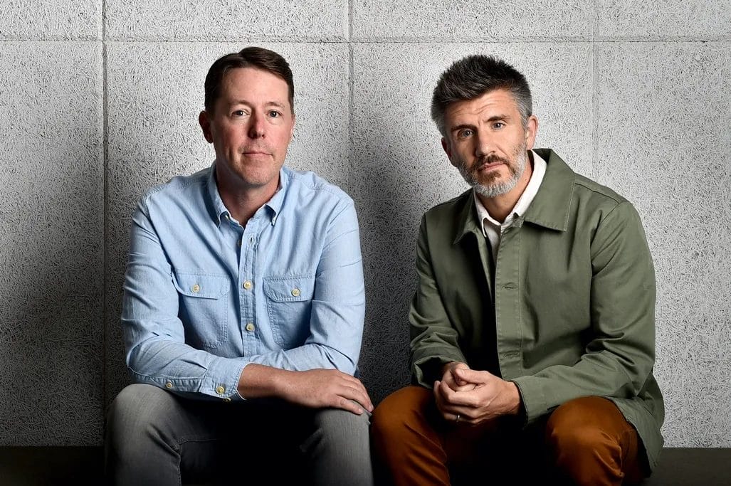 News 111 Two men sit side by side against a gray textured wall. The man on the left wears a light blue button-up shirt and gray pants, while the man on the right wears a green jacket over a white shirt and brown pants. Both look directly at the camera with neutral expressions.