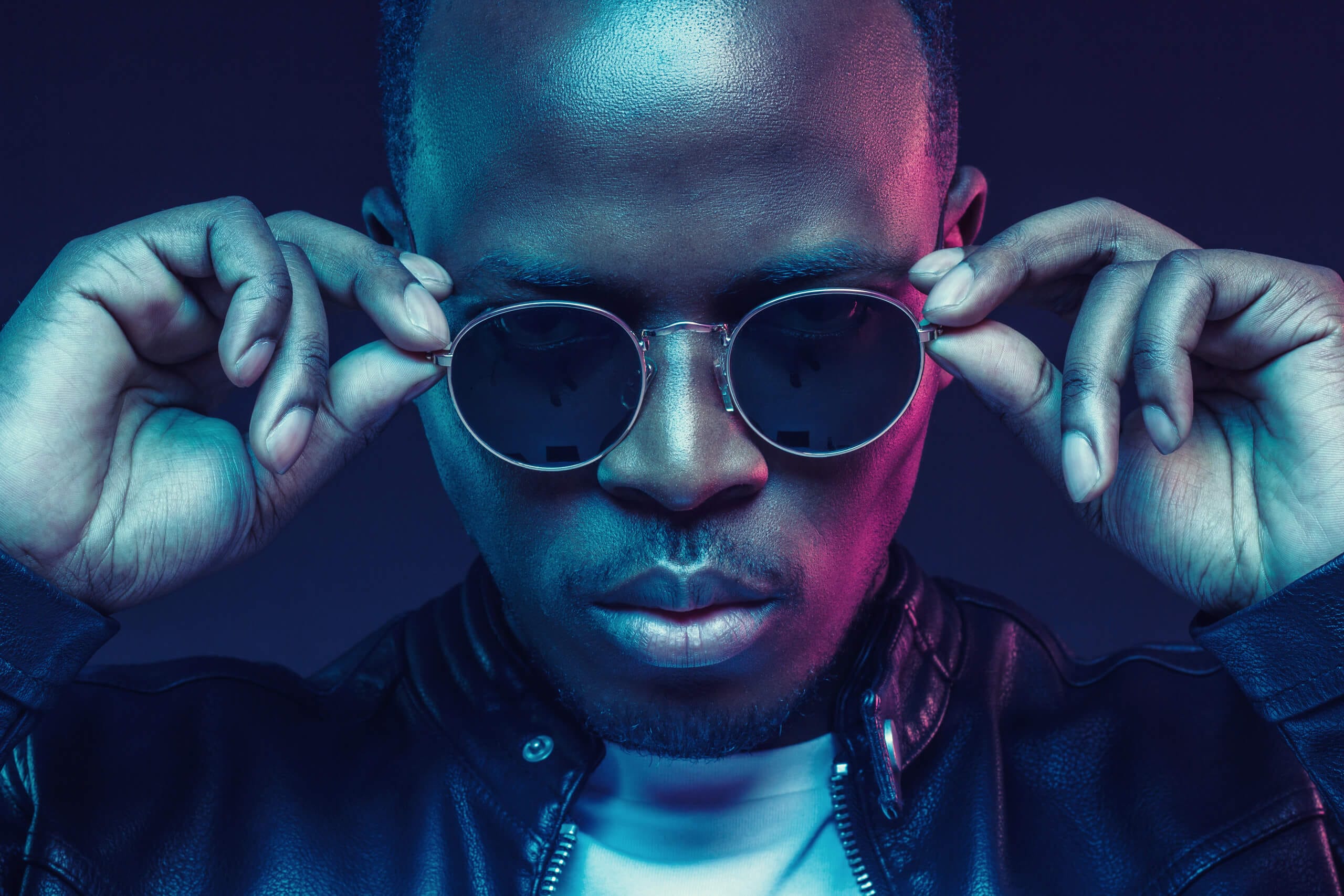 A man wearing a black leather jacket and round sunglasses adjusts his glasses with both hands. He has a serious expression and is lit by dramatic blue and pink lighting against a dark background.