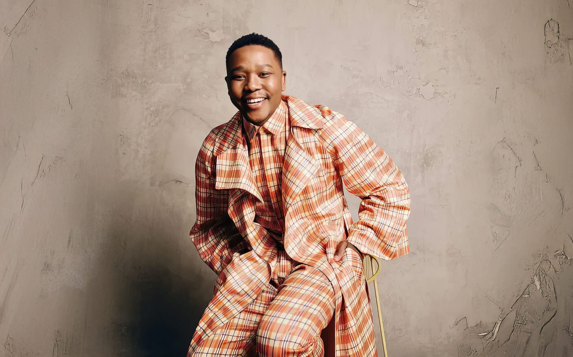 A person smiles while sitting on a stool against a textured beige wall, projecting a cheerful, relaxed demeanour reminiscent of R&B artist Langa Mavuso. They wear a matching checked outfit in orange, cream, and red, with hands resting calmly on their knees.
