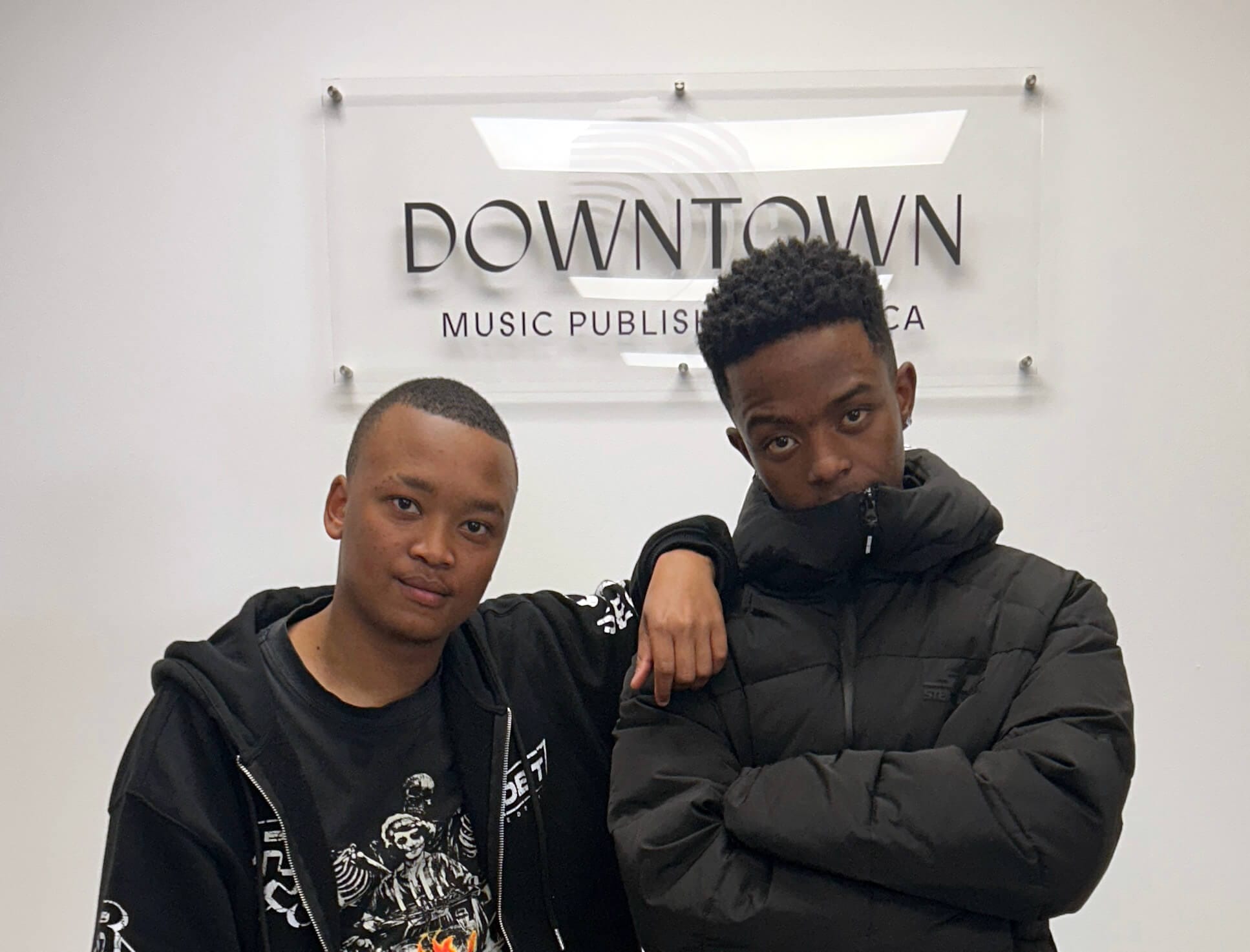 Two young men, including Rivalz, stand by a wall with a “Downtown Music Publishing Africa” sign—one in a black graphic hoodie with his arm round the other in a puffer jacket. Both look at the camera with relaxed, serious expressions.